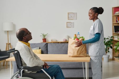 African young volunteer bringing food in paper bag for senior man in wheelchair, she smiling and talking to him in the room